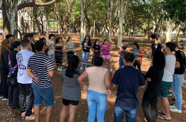 Mesmos alunos estão agora em roda, localizados em um parque, com chão de terra e há árvores ao fundo, e entre eles passa um fio de barbante, eles estão realizando alguma atividade do curso Ubunto.