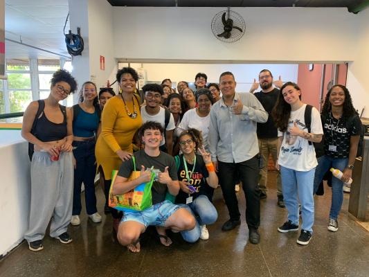 Diversos alunos posam para foto junto à Professora Débora de Souza Santos, que é uma mulher negra, de altura média, com cabelos curtos, usando um vestido amarelo e um colar étnico. Eles estão dentro de um prédio com paredes brancas e ventiladores de teto.