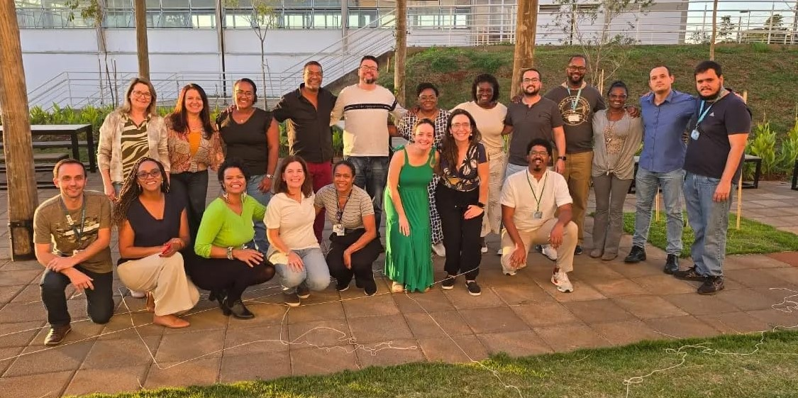 Grupo de 20 pessoas reunidas em uma área aberta, direcionados para a fotografia. Destas pessoas, 12 estão em pé e 8 abaixados em frente.