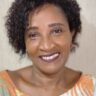 thida: Mulher negra, com cabelo curto e cacheado, ela sorri.
